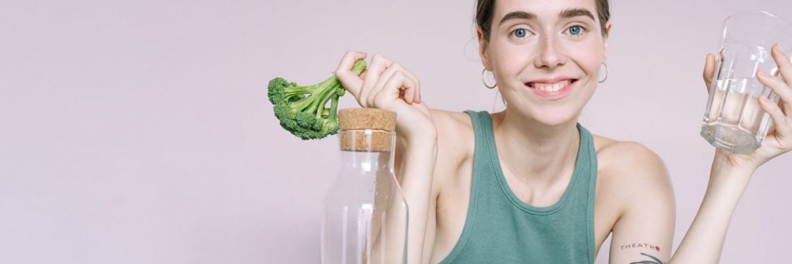 Woman in tank top smiling with broccoli, cabbage, and glass of water on table.