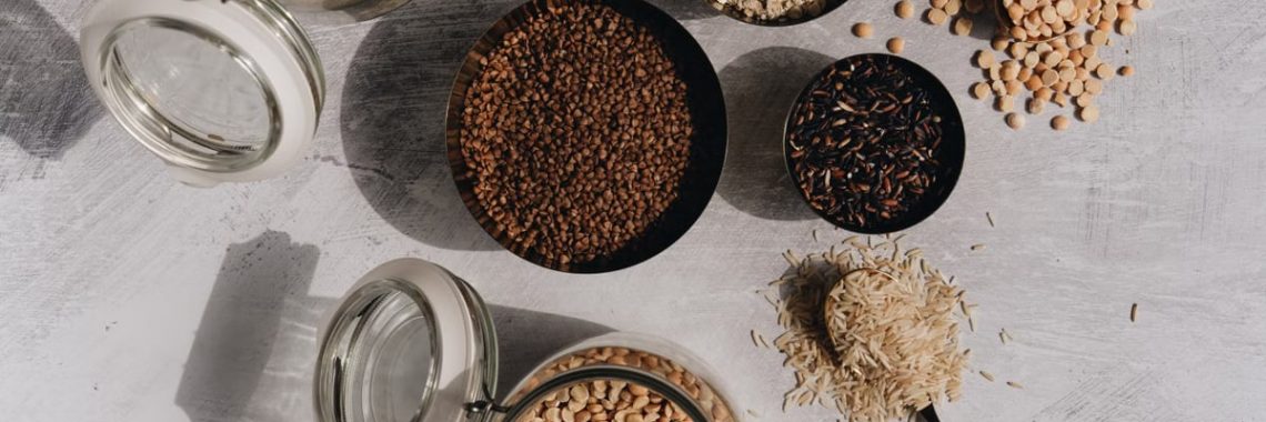 A top view of assorted grains and pulses in jars and bowls on a textured surface with shadow play.